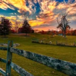Scenic autumn landscape at Watson Estate, Deer Creek Winery, featuring a wooden fence in the foreground, gently rolling grassy hills, and trees with red and orange fall foliage. A striking sunset fills the sky with vibrant swirls of orange, pink, and purple clouds against a deep blue backdrop.