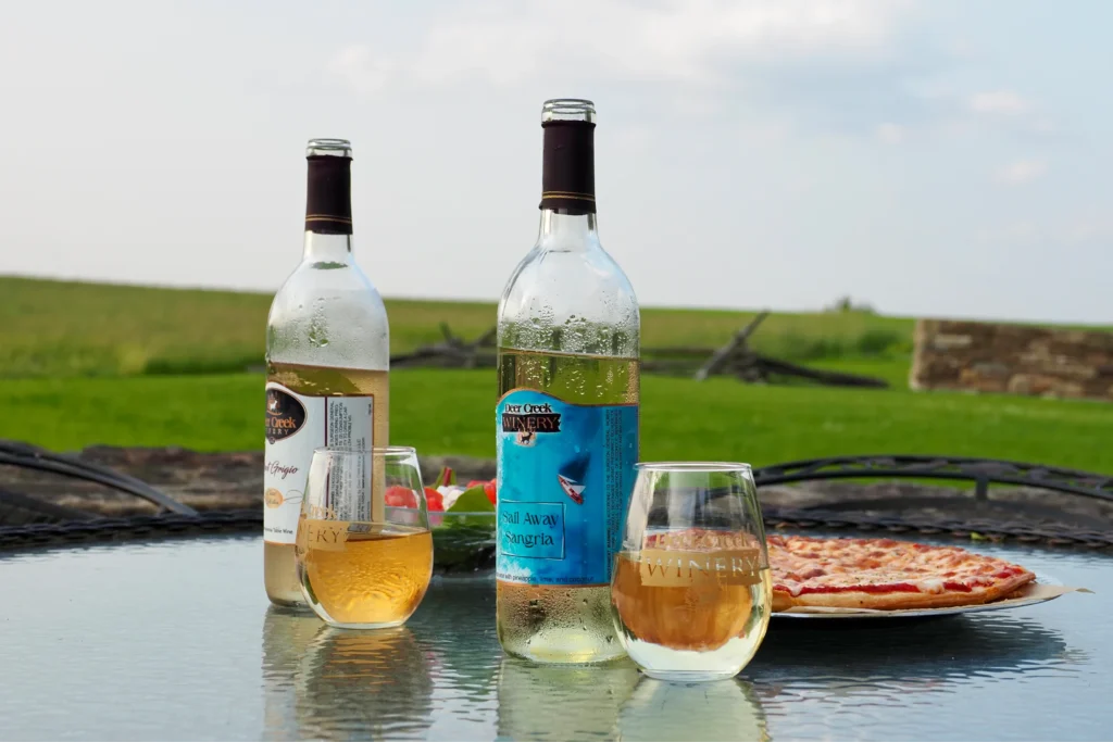 Two chilled bottles of Deer Creek Winery wine, including Sail Away Sangria, sit on a glass table alongside two branded wine glasses and a fresh pizza, with the scenic countryside of Watson Estate at Deer Creek Winery in the background.
