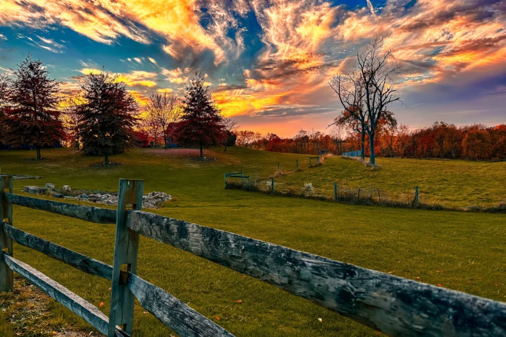 Scenic autumn landscape at Watson Estate, Deer Creek Winery, featuring a wooden fence in the foreground, gently rolling grassy hills, and trees with red and orange fall foliage. A striking sunset fills the sky with vibrant swirls of orange, pink, and purple clouds against a deep blue backdrop.