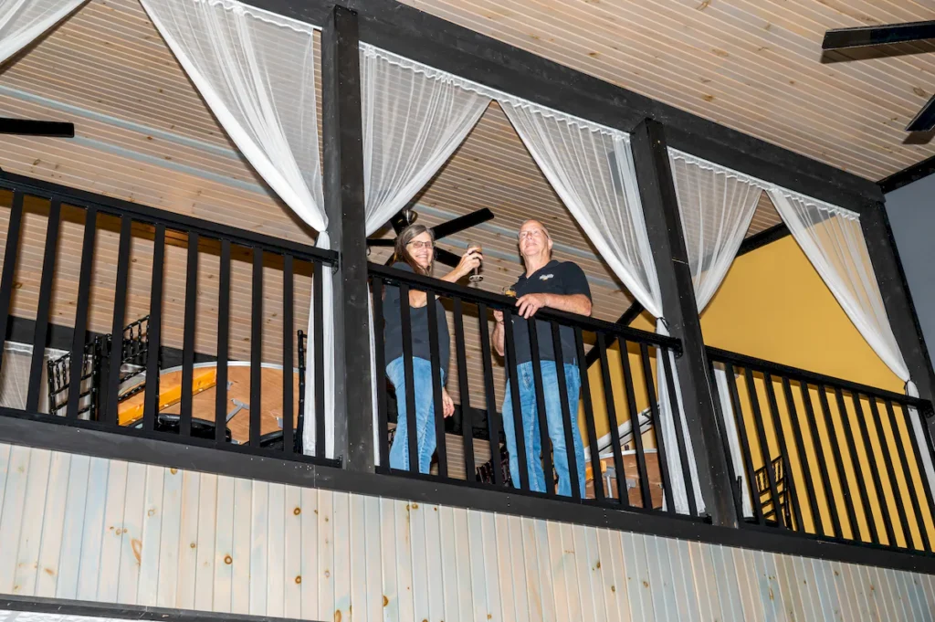 Denis and Rhonda Brooks standing in the upper Mezzanine area against railing drinking wine at The Gathering Place at The Inn Event Center at Deer Creek Winery at Brooks Estate